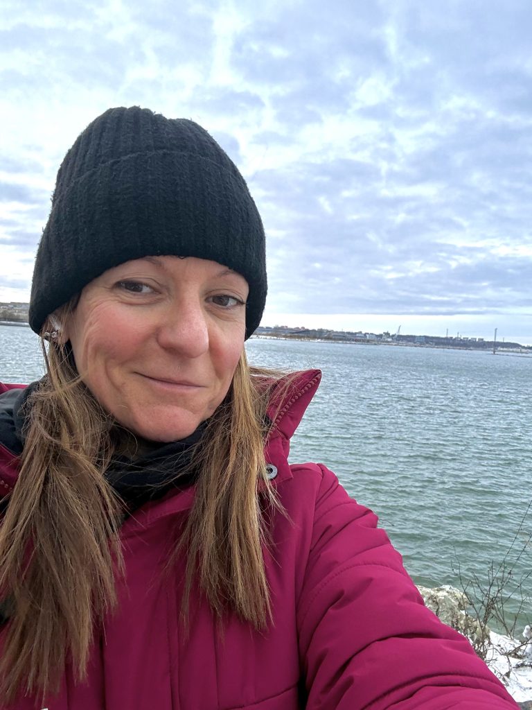 A woman wearing a black beanie and a pink jacket takes a selfie by the water on a cloudy day.